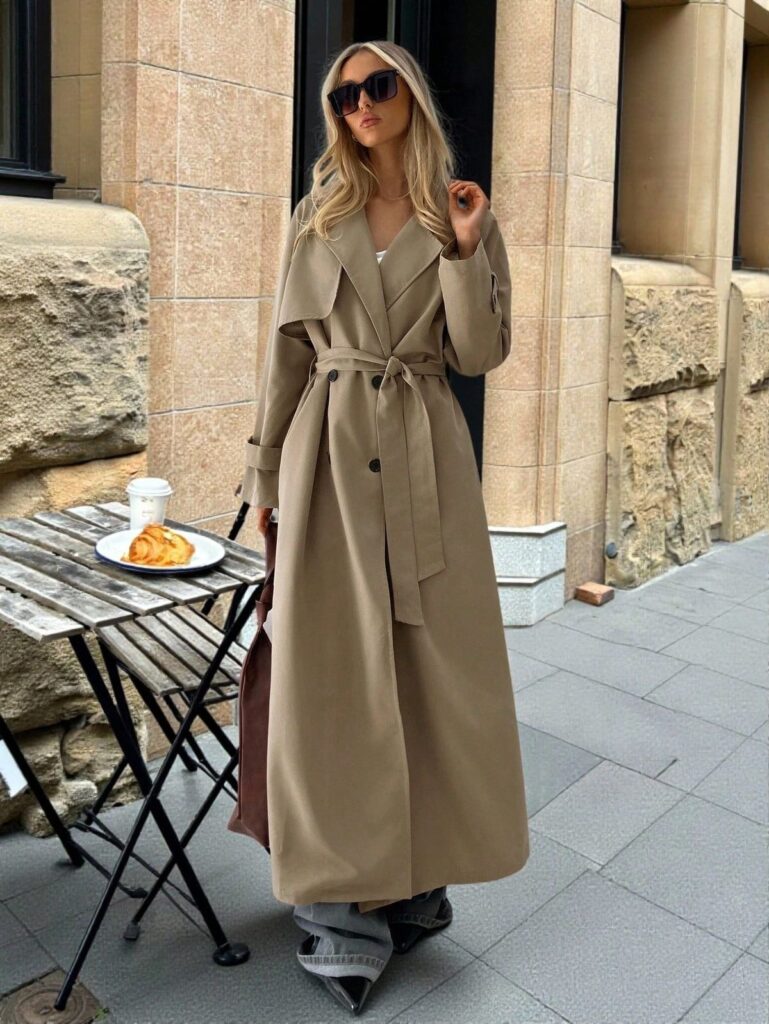 Un trench long ceinturé jusqu’aux chevilles porté sur un jean gris clair large : un look automne-hiver élégant et tendance.