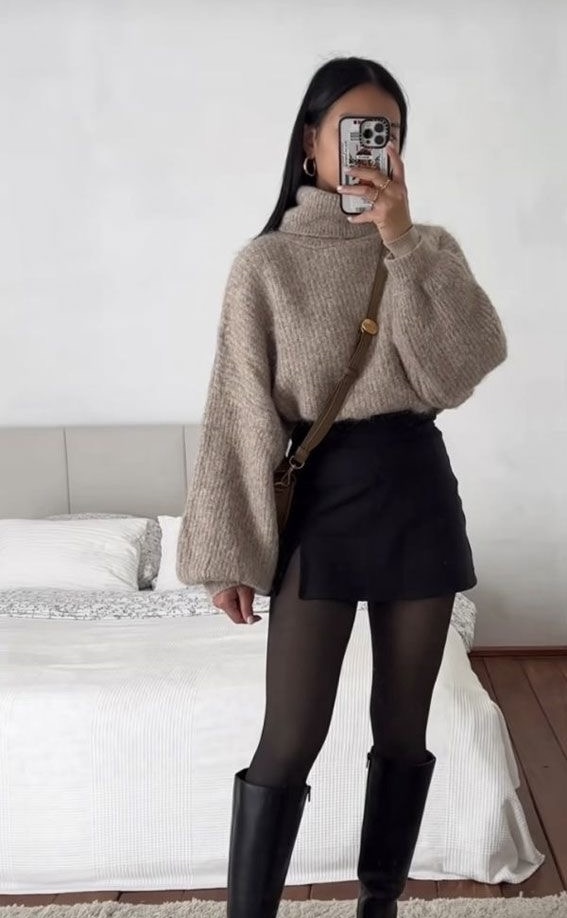 Un pull oversize beige en maille à col roulé, associé à une jupe-short noire et des bottes hautes noires. Un look minimaliste et élégant, parfait pour la mi-saison.
