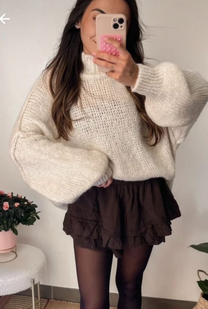 Un pull oversize beige en maille à col roulé, associé à une jupe-short noire et des bottes hautes noires. Un look minimaliste et élégant, parfait pour la mi-saison.